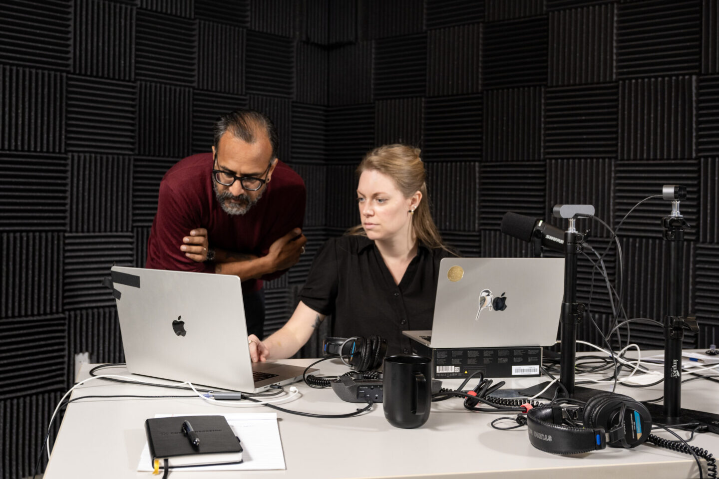 Two people work together in a soundproof room with foam panels. They are using laptops and audio equipment on a white table, appearing focused and engaged in their task.