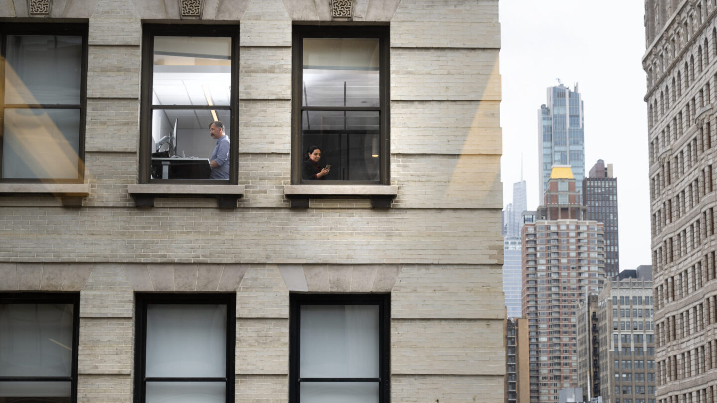 Two people are seen through office windows in a beige city building; one is standing and the other is seated. Tall skyscrapers and green trees are visible in the background under a cloudy sky.