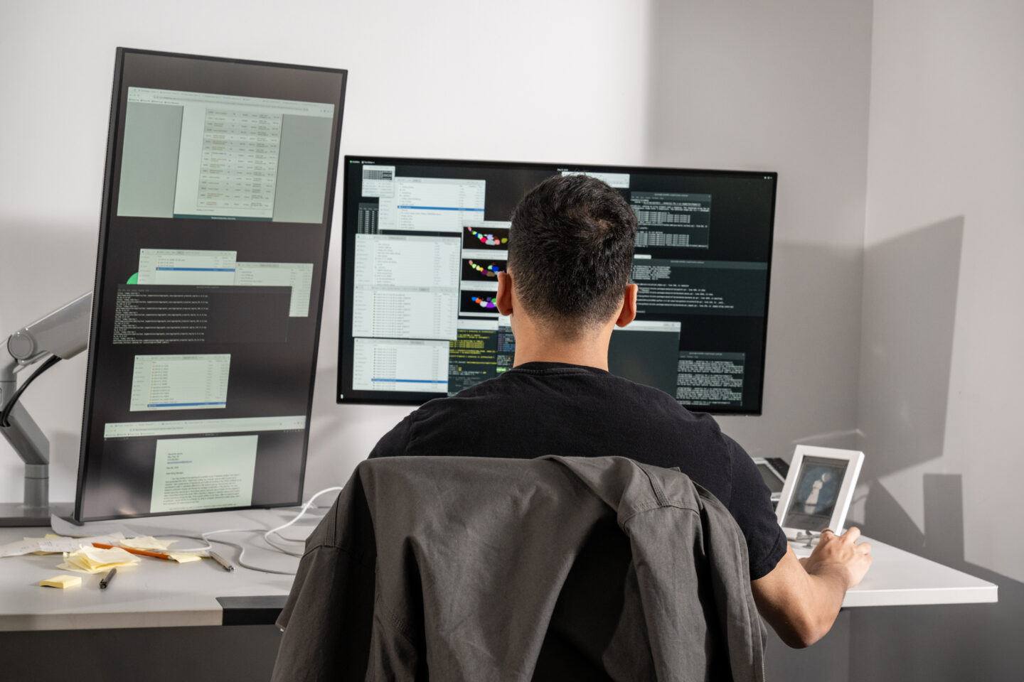A person sits at a desk with three large computer monitors displaying code and data, working in an office setting. A jacket hangs on the chair, and sticky notes are on the desk. The person is viewed from behind.