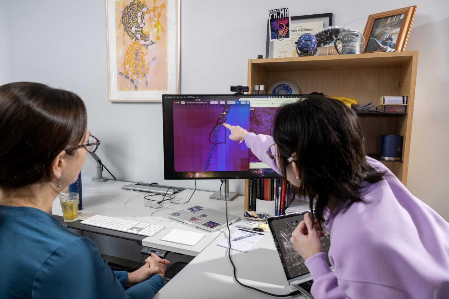 Two people sit at a desk, looking at a computer monitor. One person points at a screen displaying colorful data and a graph, while the other observes. Books, art, and awards are visible in the background.