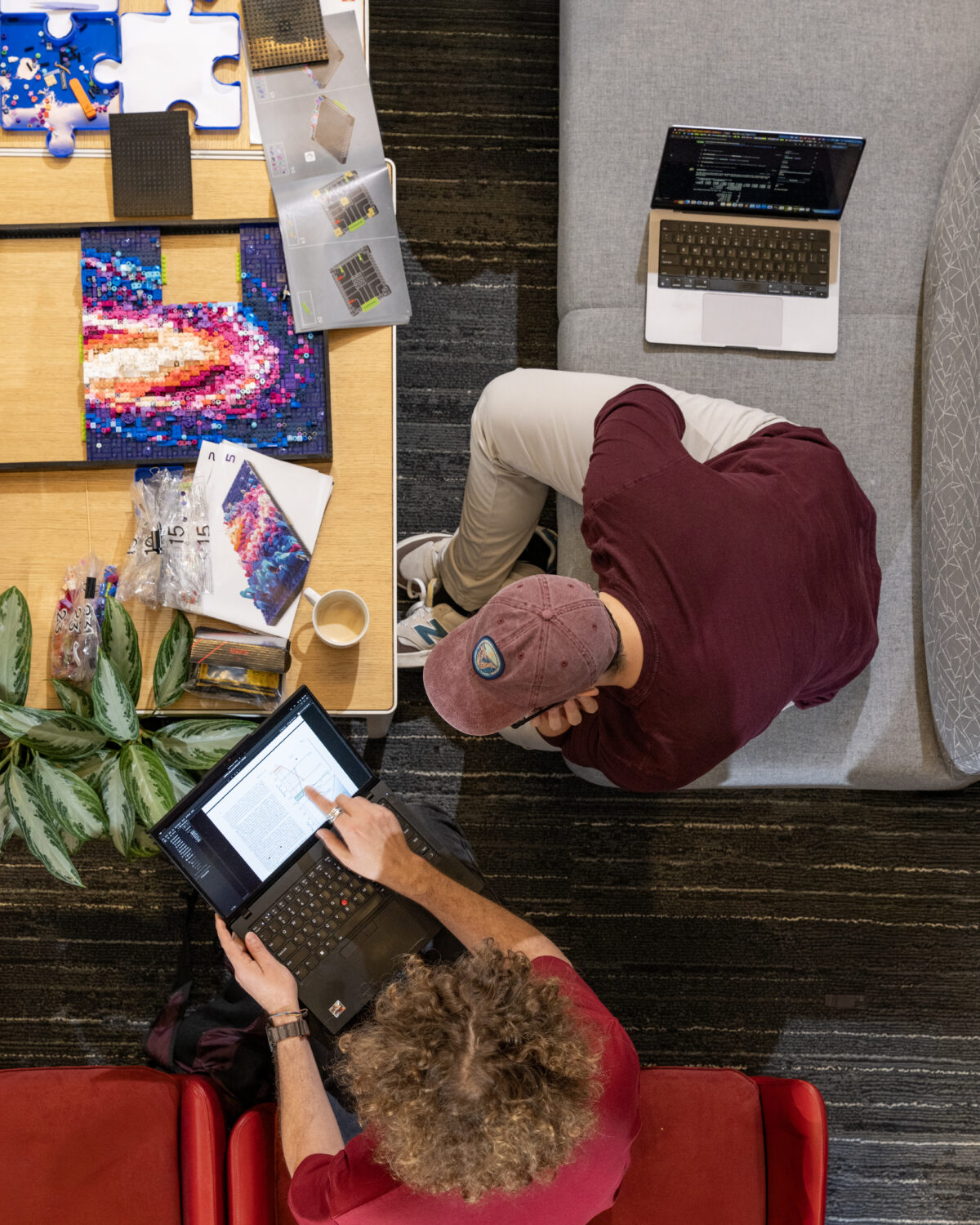 Two people work on laptops at a table with puzzle pieces, papers, a coffee cup, and a plant. The photo is taken from above, showing a collaborative and creative workspace setting.