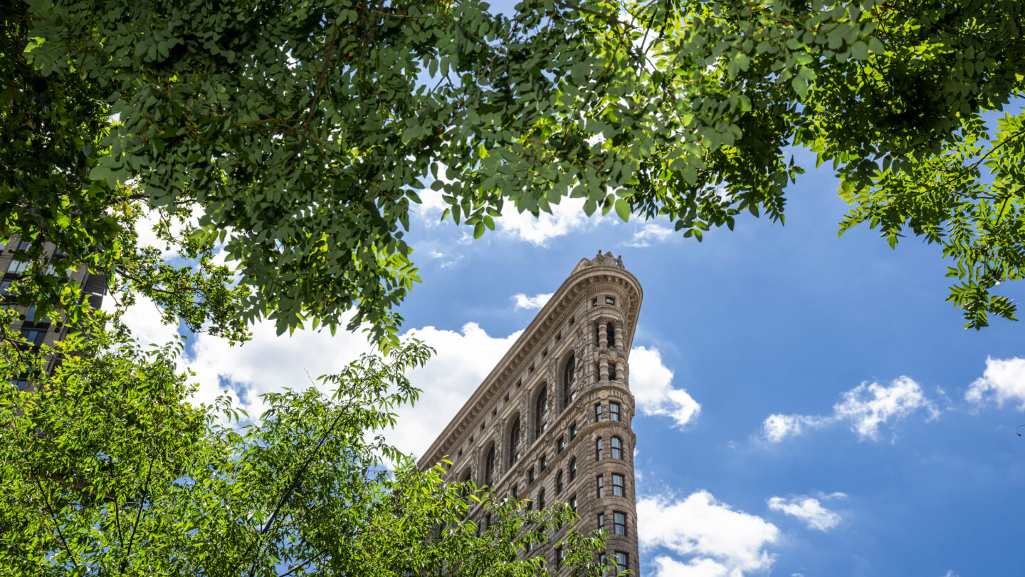 A tall, triangular building seen from below, framed by green tree branches, under a bright blue sky with scattered white clouds.