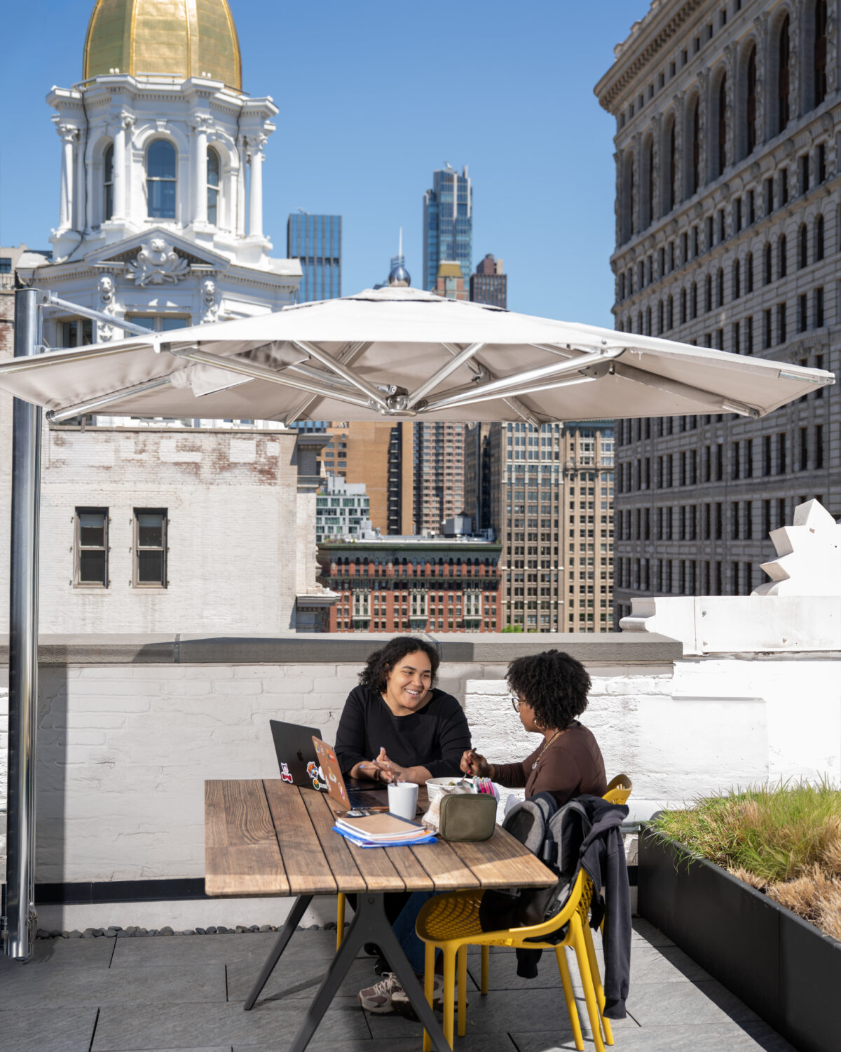 Two people sit at a table on a rooftop patio under an umbrella, talking and working on laptops, with city buildings and a golden-domed structure in the background.