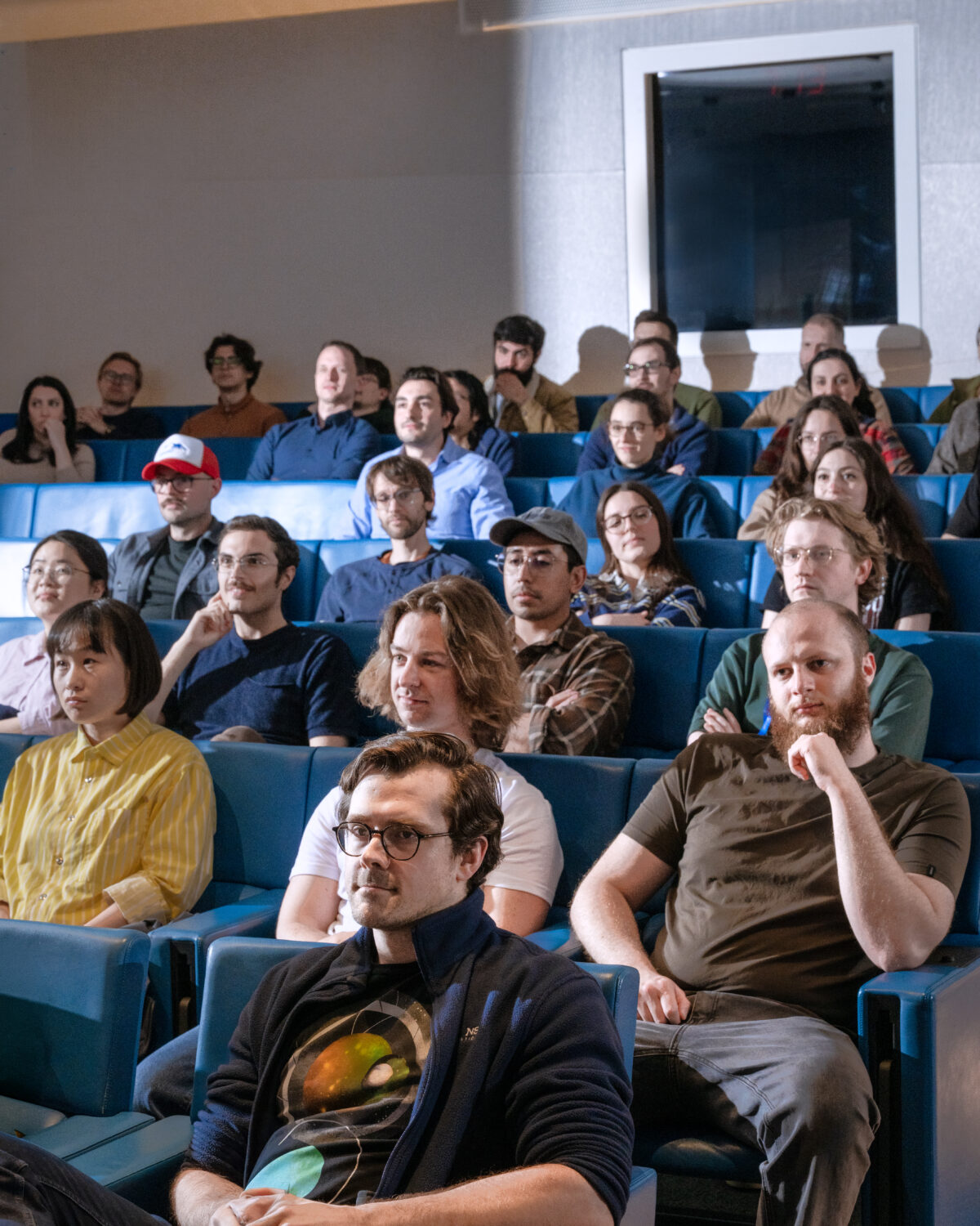 A diverse group of people sit in blue auditorium seats, attentively watching something off camera, some appearing thoughtful, others neutral or smiling.