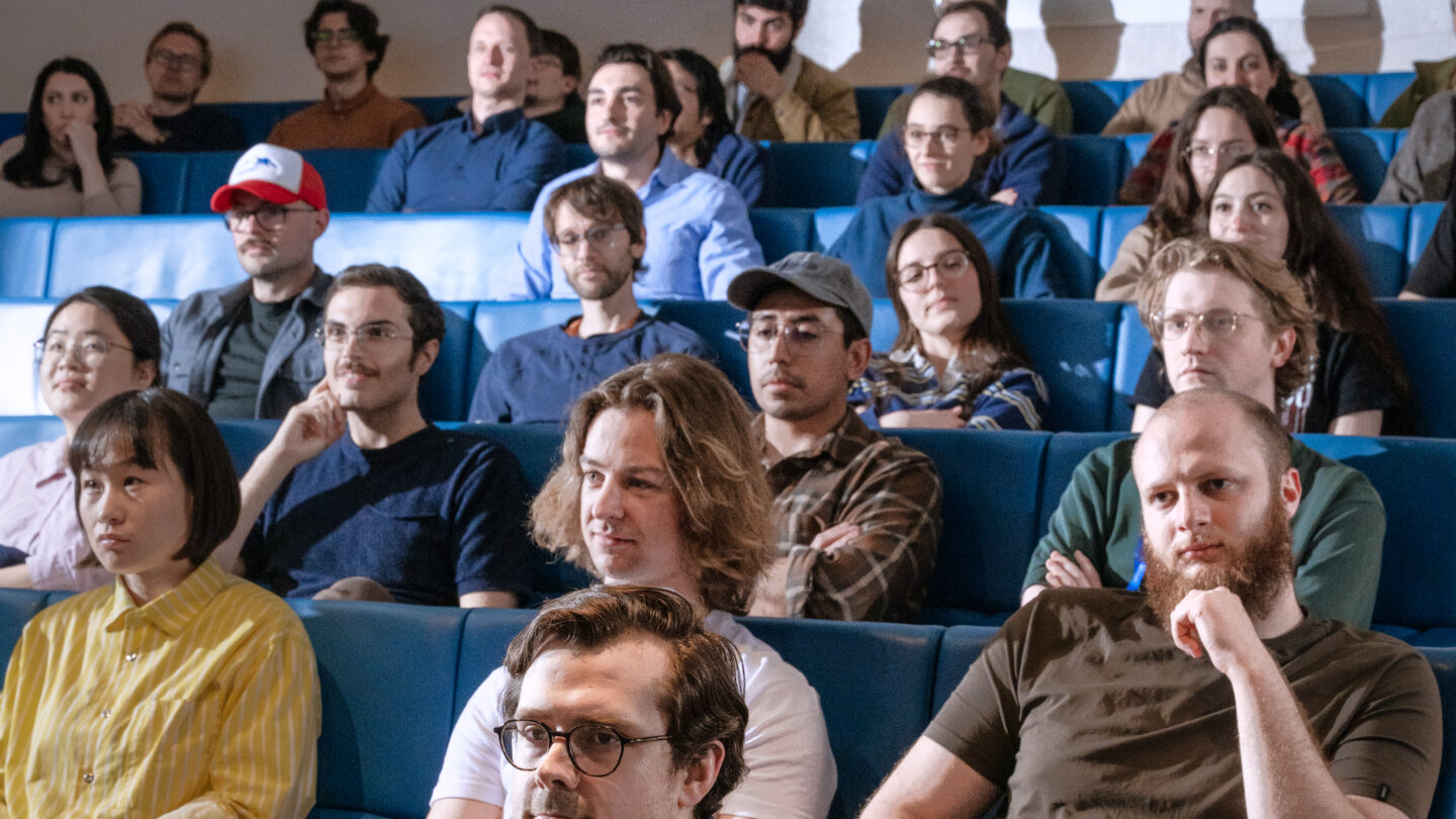 A diverse group of people sit in blue auditorium seats, attentively watching something off camera, some appearing thoughtful, others neutral or smiling.