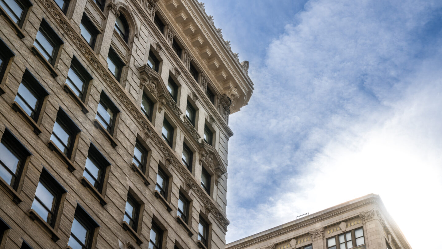 Two historic stone buildings with many windows rise against a bright blue sky with wispy clouds, sunlight shining from the lower right corner of the image.
