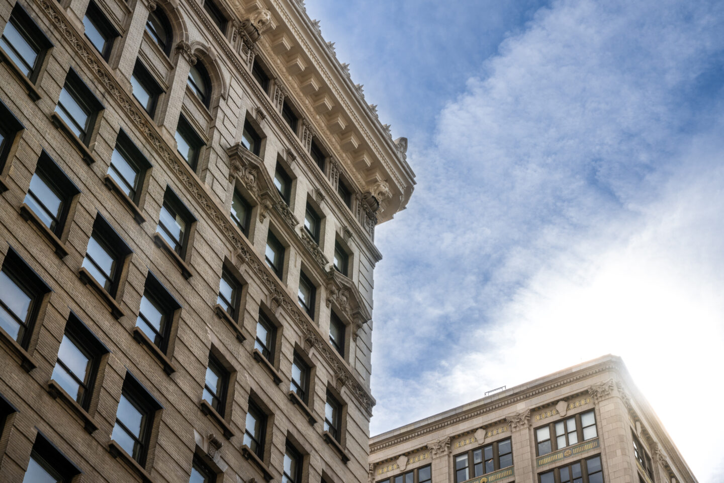 Two historic stone buildings with many windows rise against a bright blue sky with wispy clouds, sunlight shining from the lower right corner of the image.