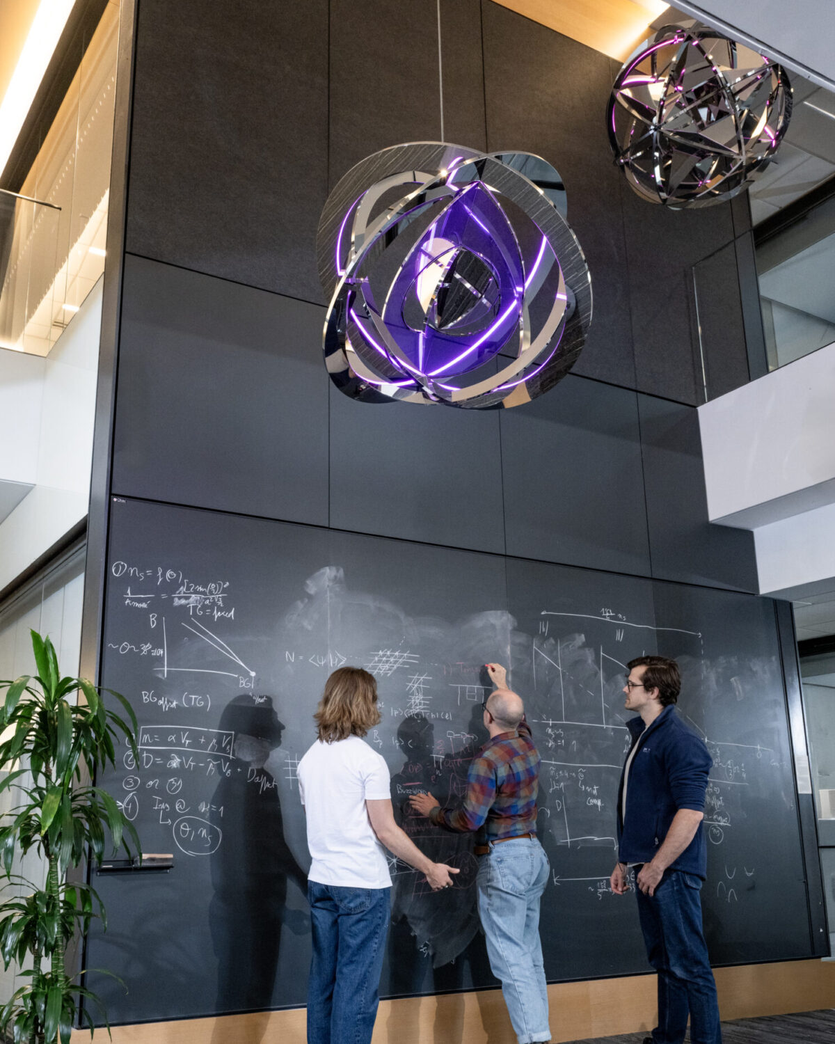 Three people stand at a large blackboard covered with mathematical equations in a modern office, discussing and pointing at the board under a futuristic hanging light fixture.