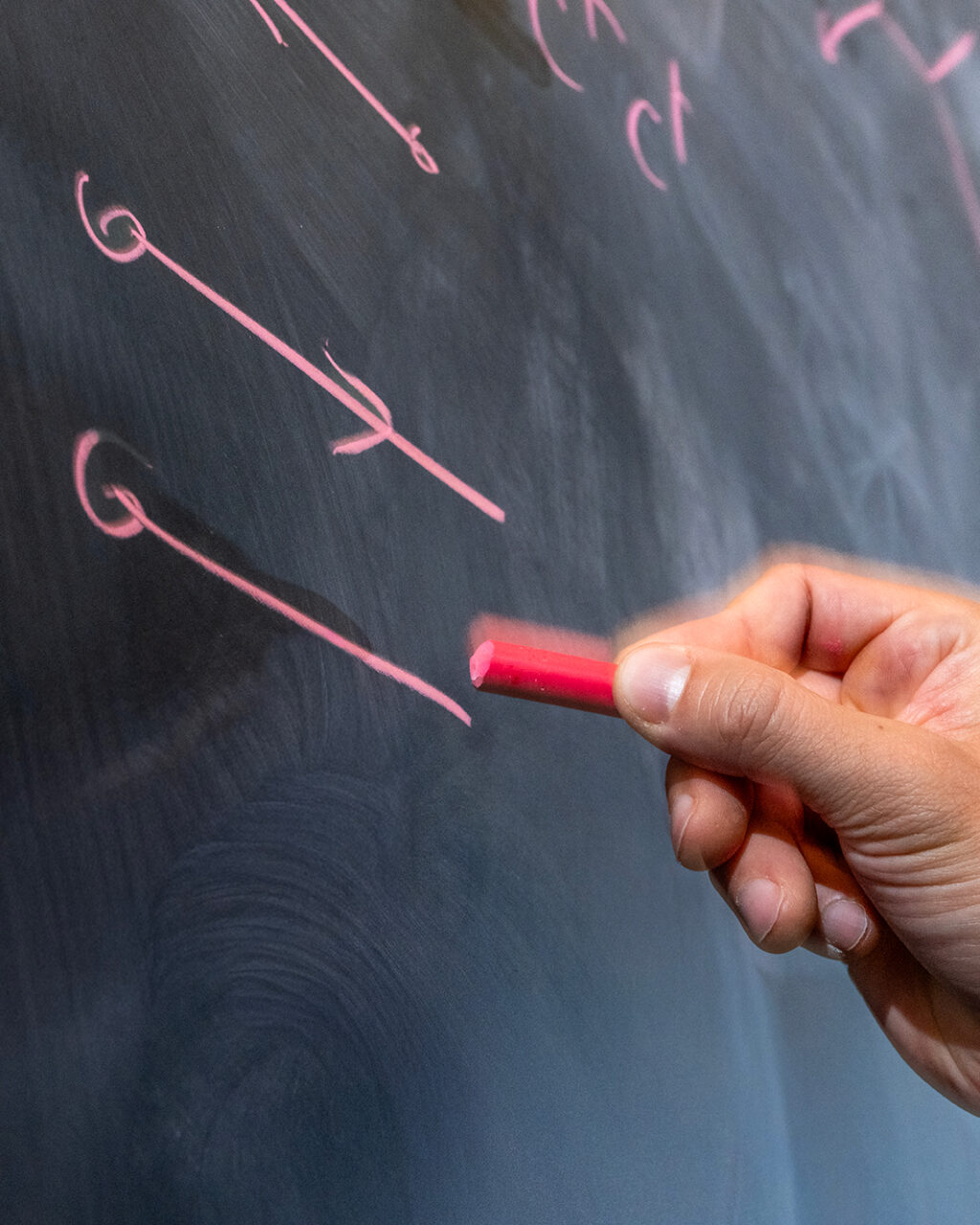 A hand holds pink chalk while drawing arrows and symbols on a blackboard.