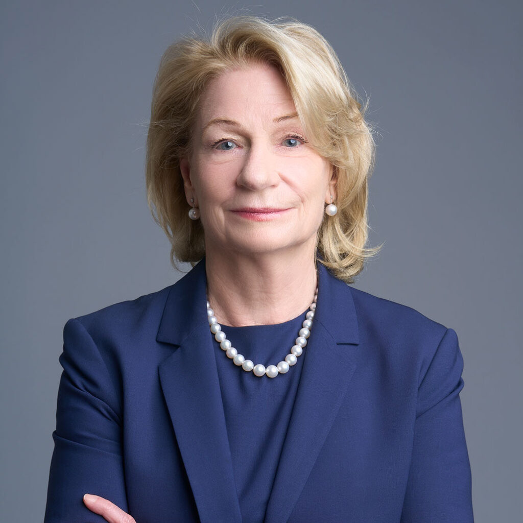 Confident older woman with blond hair wearing a navy blue suit and pearl necklace, standing with arms crossed against a plain gray background.