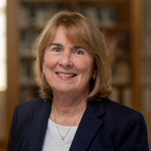 A woman with light brown hair, wearing a navy blazer and white shirt, smiles at the camera. She is in an indoor setting with blurred bookshelves in the background.
