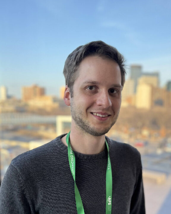 A man wearing a grey sweater and a green conference lanyard smiles at the camera, with a city skyline visible through the window behind him.