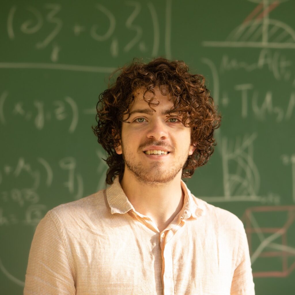 A person with curly hair and a light shirt stands smiling in front of a chalkboard filled with mathematical equations and geometric diagrams.