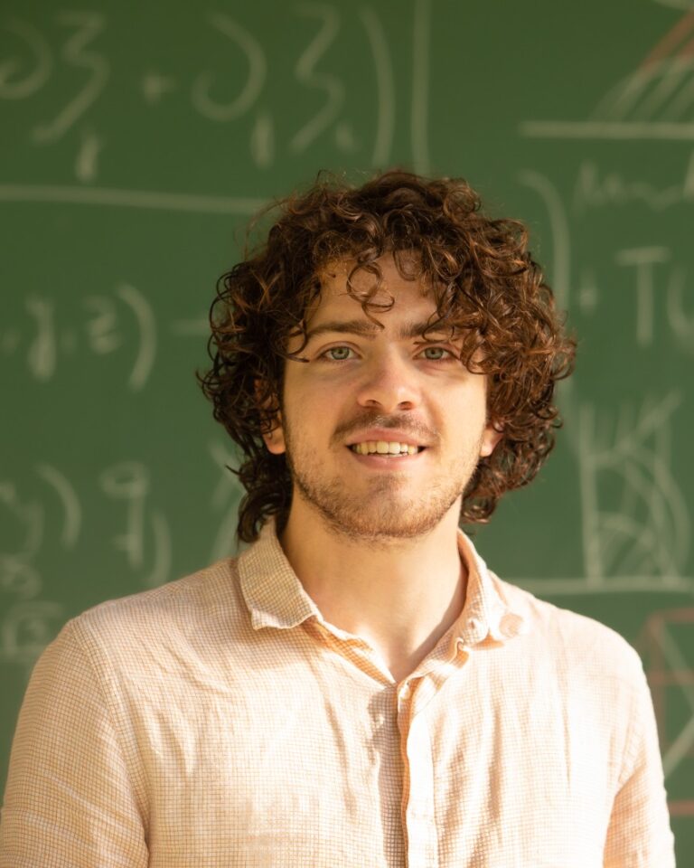 A person with curly hair and a light shirt stands smiling in front of a chalkboard filled with mathematical equations and geometric diagrams.