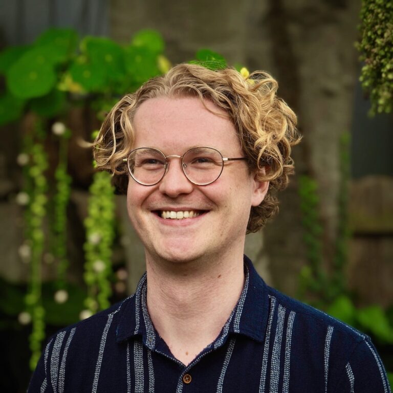 A smiling person with curly blond hair and glasses stands outdoors in front of green plants, wearing a dark blue striped shirt.
