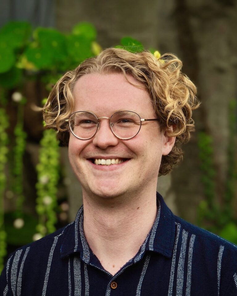 A smiling person with curly blond hair and glasses stands outdoors in front of green plants, wearing a dark blue striped shirt.