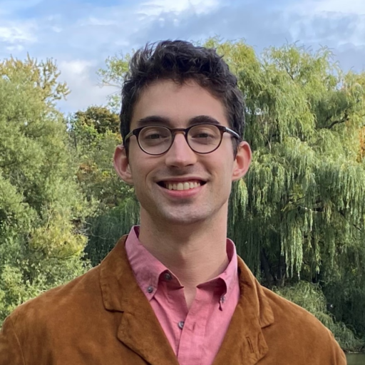 A person with short brown hair and glasses smiles outdoors, wearing a brown jacket over a pink shirt. Lush green trees are in the background under a partly cloudy sky.