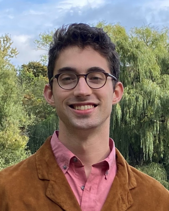 A person with short brown hair and glasses smiles outdoors, wearing a brown jacket over a pink shirt. Lush green trees are in the background under a partly cloudy sky.