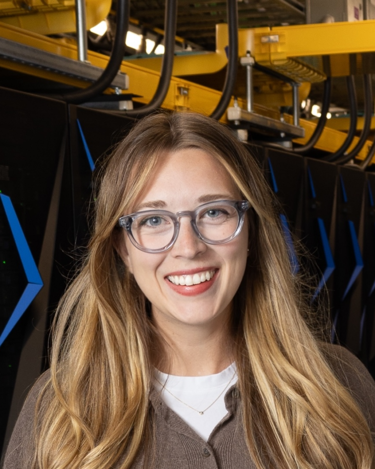 A woman with long blonde hair and glasses smiles in front of rows of black computer servers with blue accents and yellow cable trays overhead.