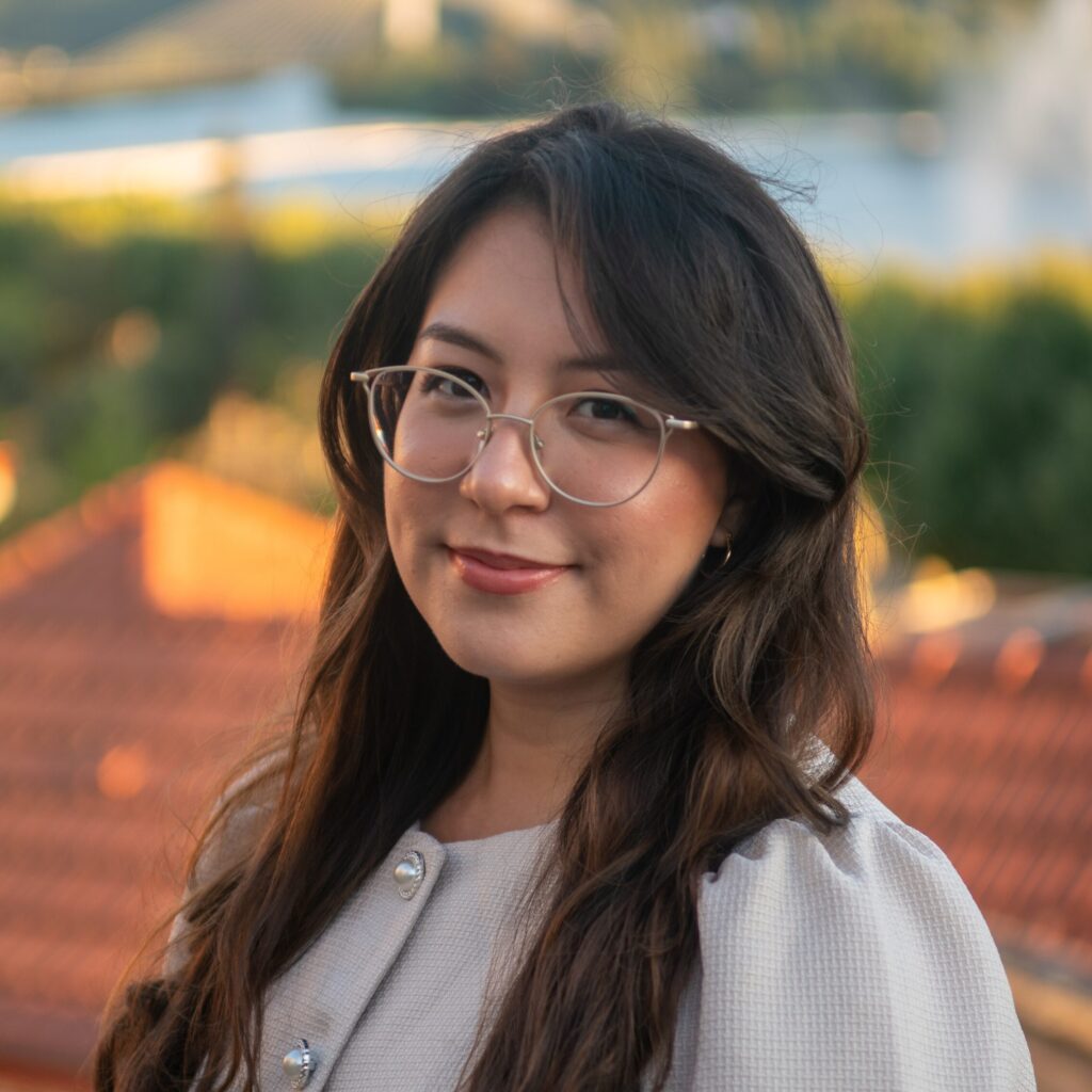 A woman with long brown hair and glasses smiles outdoors with a blurred background of rooftops, greenery, and a river.