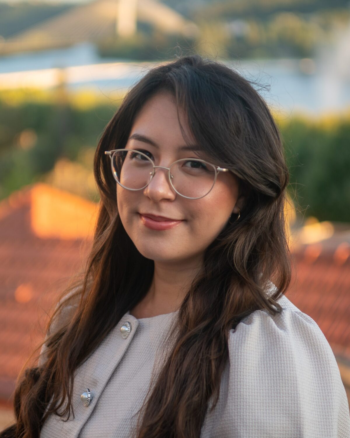 A woman with long brown hair and glasses smiles outdoors with a blurred background of rooftops, greenery, and a river.