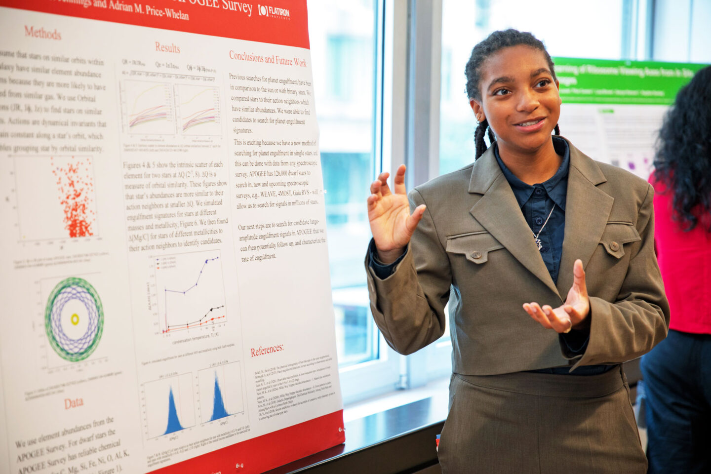 A young woman in a gray suit jacket gestures while presenting a scientific research poster about planet engulfment.