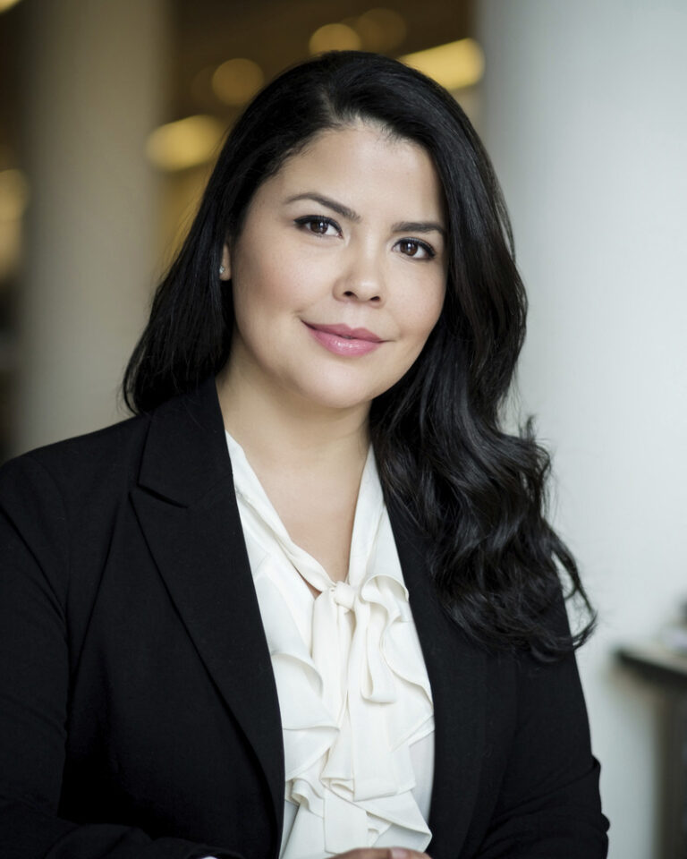 A professional headshot of a woman with long dark hair wearing a white blouse and a black blazer.