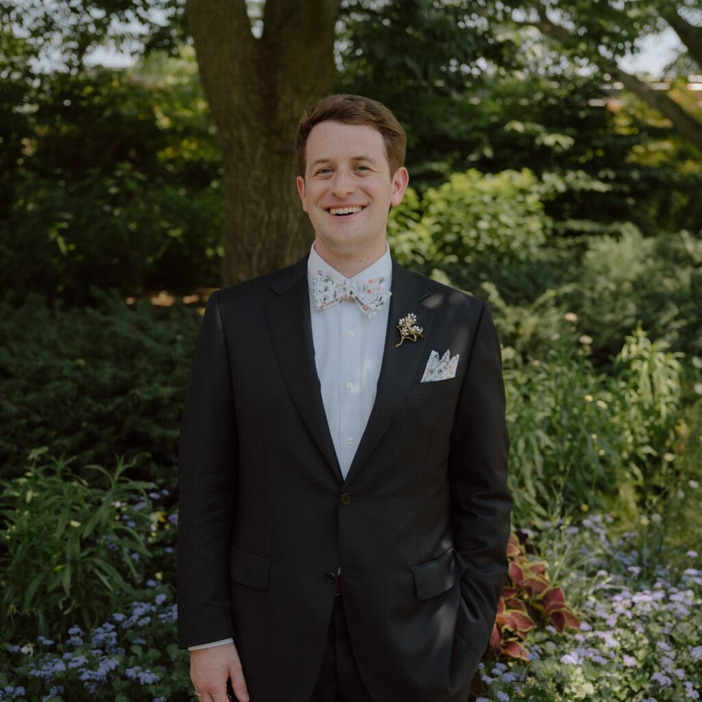 A smiling man in a dark suit, white dress shirt, and floral bow tie stands in a garden.