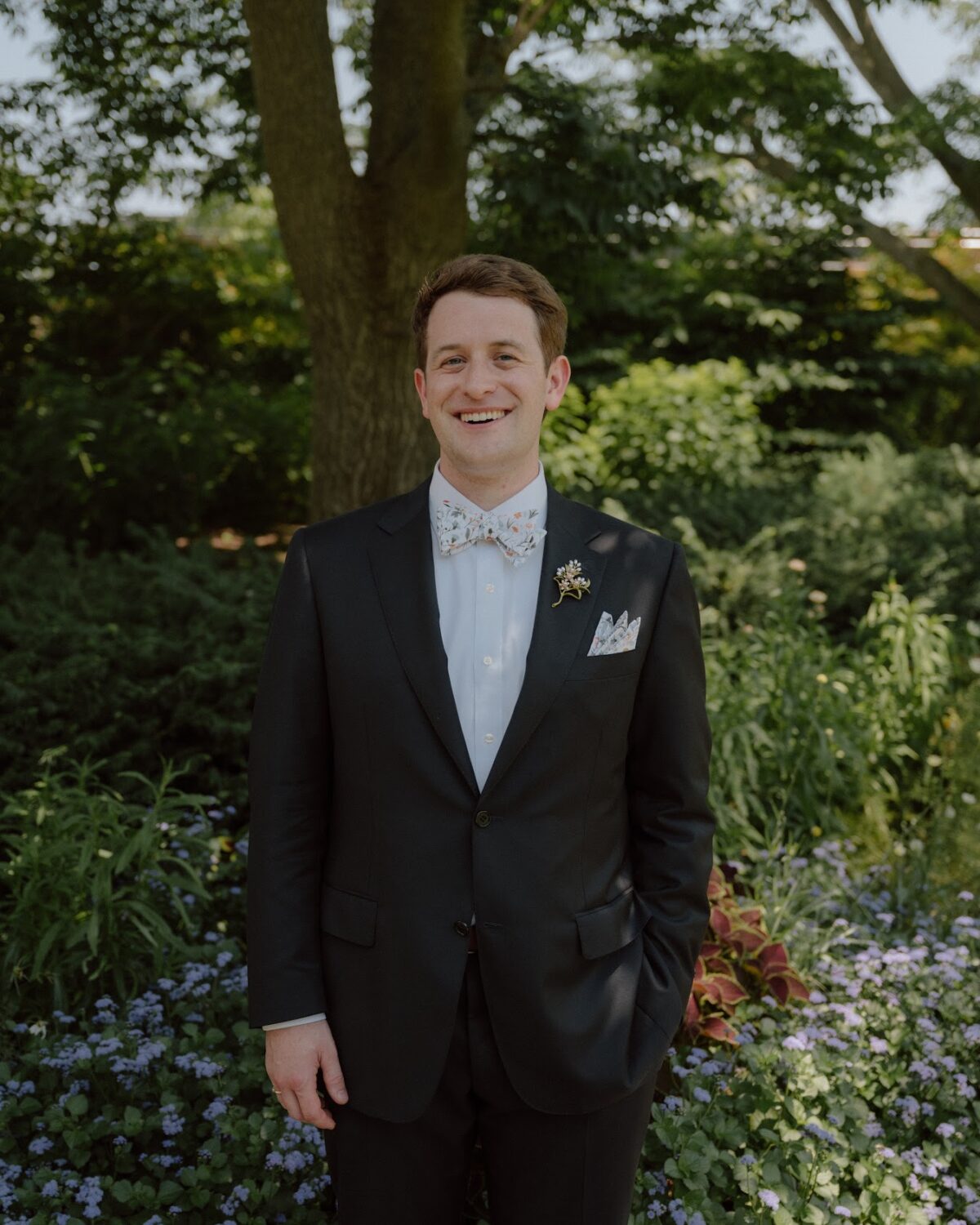 A smiling man in a dark suit, white dress shirt, and floral bow tie stands in a garden.