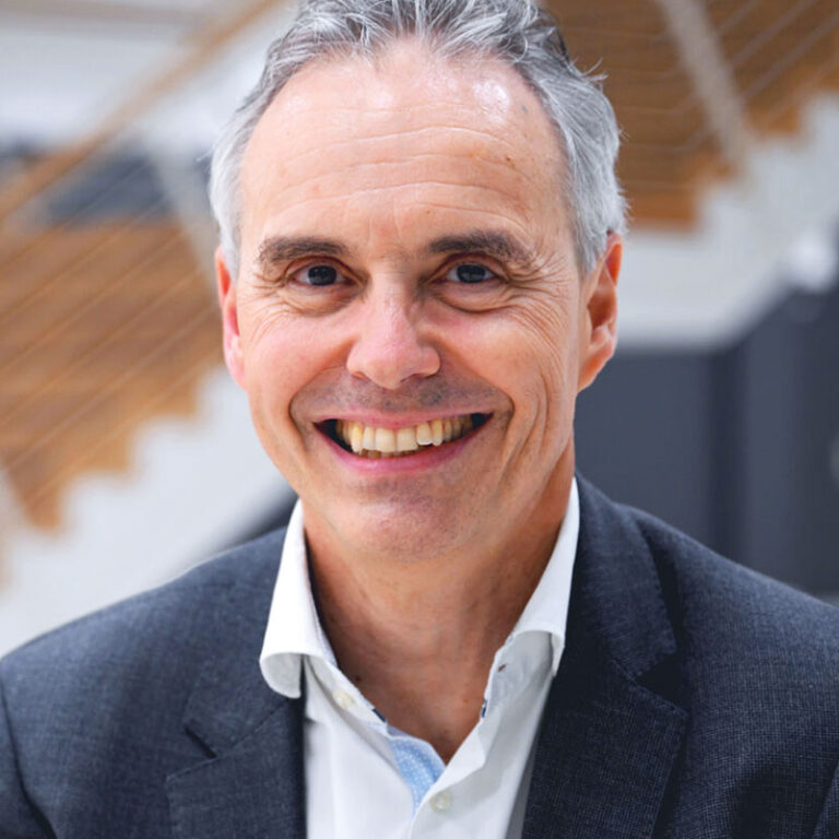 A smiling middle-aged man with gray hair wearing a dark blazer and white dress shirt.