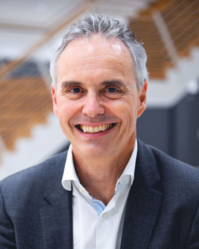A smiling middle-aged man with gray hair wearing a dark blazer and white dress shirt.