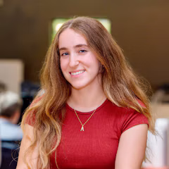 A smiling young woman with long wavy hair wears a red top and a small letter A pendant necklace.