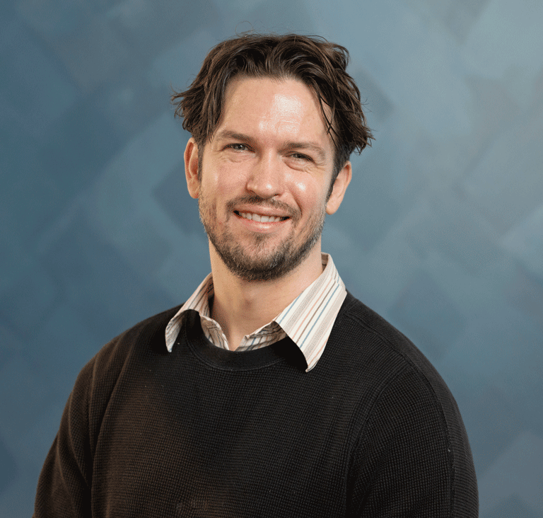 A smiling man with a light beard wearing a dark sweater over a collared shirt.