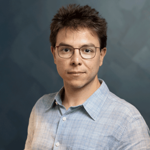 A man wearing glasses and a light blue button-down shirt looks toward the camera.