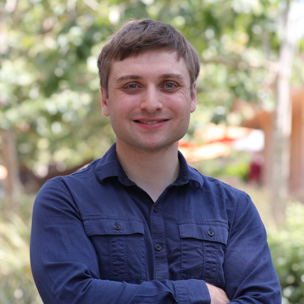 A man in a blue button-down shirt smiles with his arms crossed outdoors.