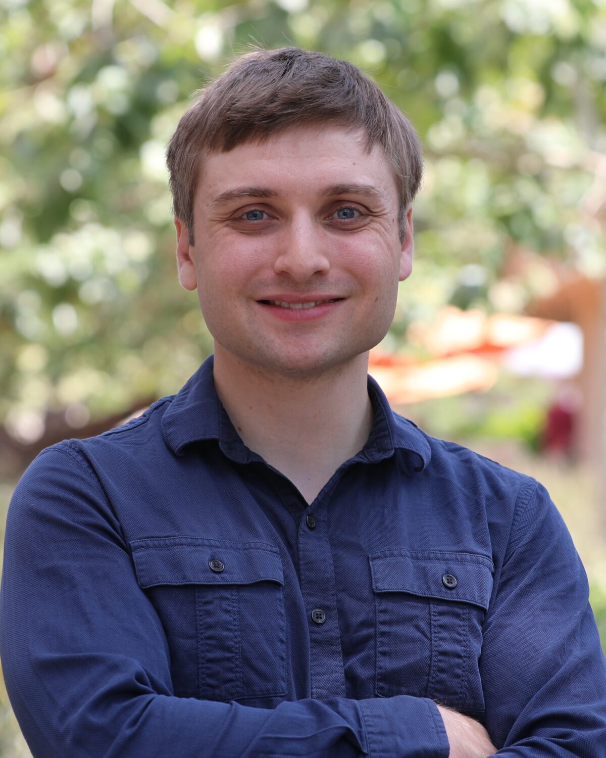 A man in a blue button-down shirt smiles with his arms crossed outdoors.