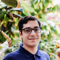 A man with dark hair and glasses wearing a blue polo shirt, positioned in front of green foliage.