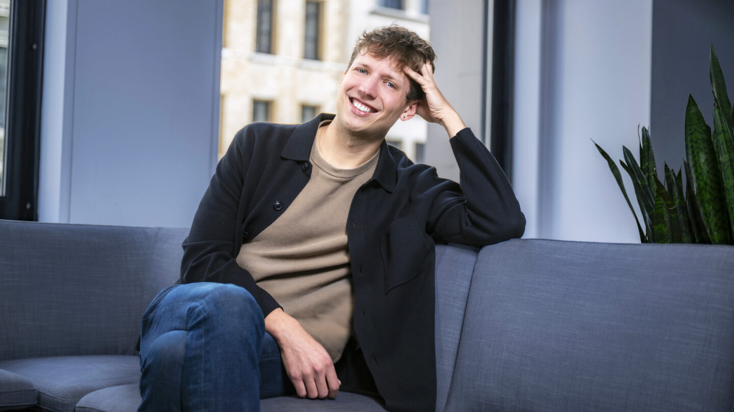 A smiling young man with his hand to his head, sitting on a gray sofa.