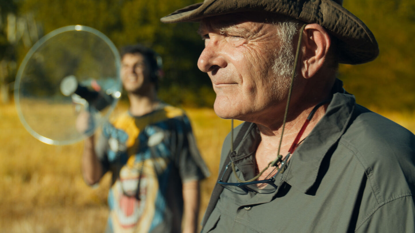 An older man wearing a sun hat stands in a field while a person in the blurry background holds a parabolic microphone.