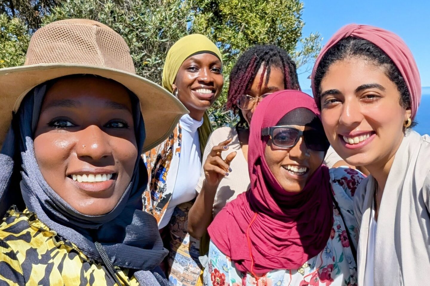 Five smiling women pose for a selfie outdoors with a clear blue sky and ocean in the background.