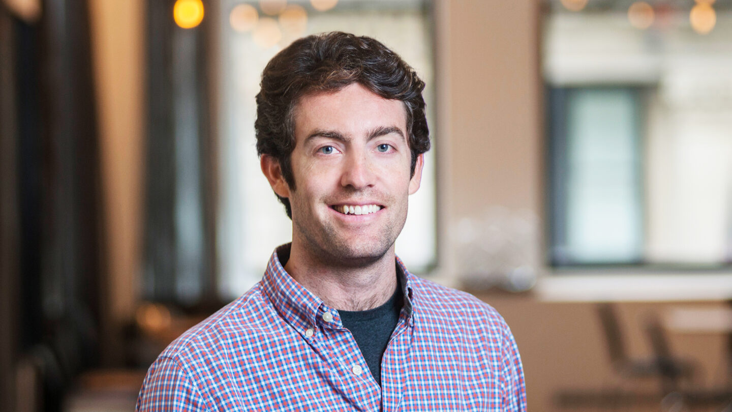A man with dark hair smiling at the camera, wearing a blue and red plaid button-down shirt.