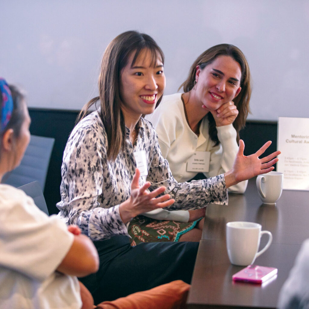 A woman speaks to a small group at a table during a meeting with a sign labeled Mentoring and Cultural Awareness nearby.