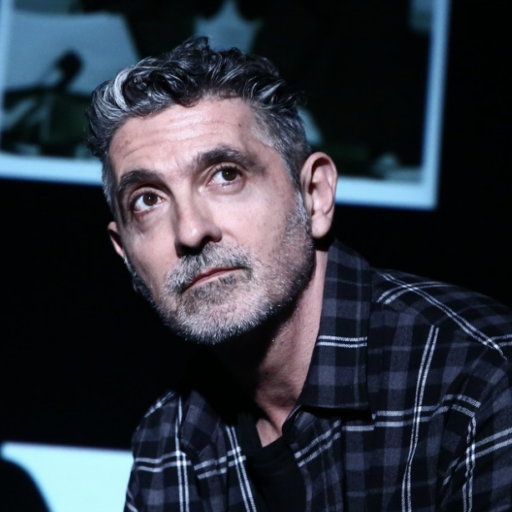 A man with graying dark hair and a beard wearing a black and gray plaid shirt.