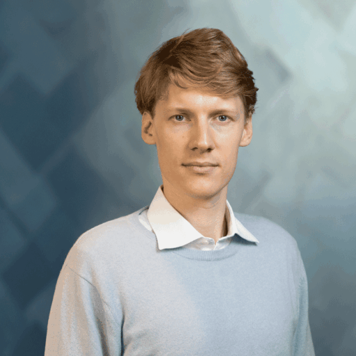 A young man with light brown hair wearing a light blue crew neck sweater over a white collared shirt.