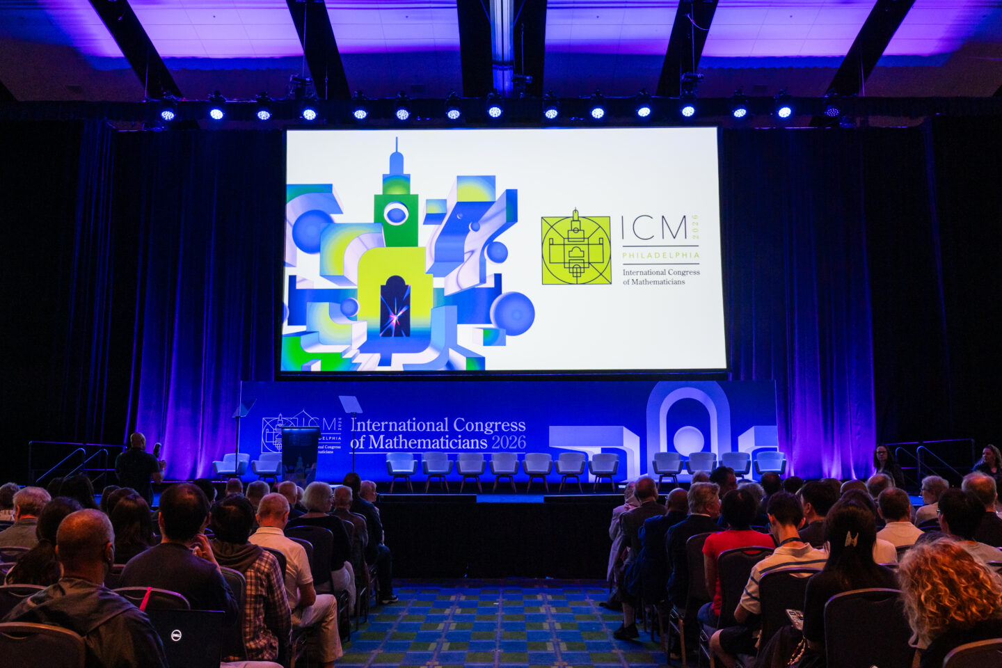 A large screen at a conference displays the International Congress of Mathematicians 2026 logo before an audience in a darkened room.