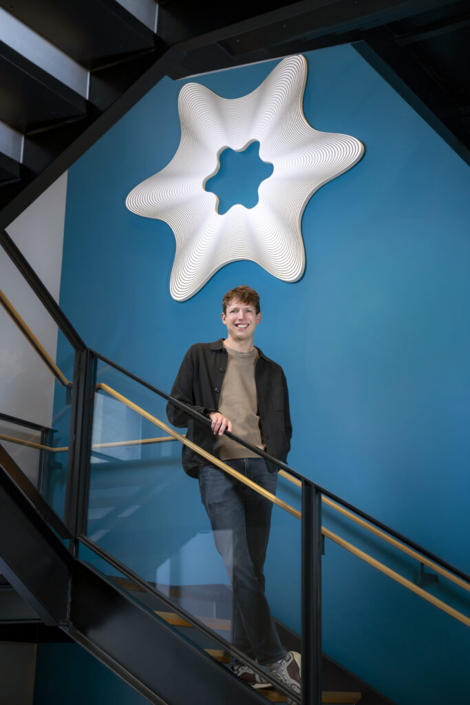 A smiling young man stands on a staircase in front of a blue wall featuring a large, white, star-shaped sculptural element.