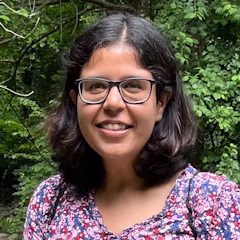 A person with shoulder-length dark hair and glasses smiling in front of green foliage.