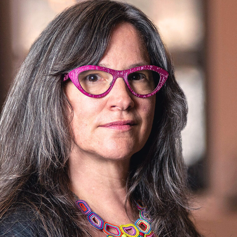 A woman with long graying hair wearing bright pink geometric glasses and a colorful beaded statement necklace.