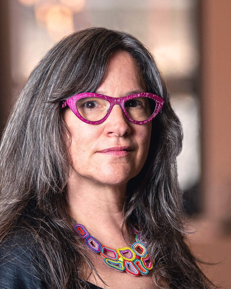 A woman with long graying hair wearing bright pink geometric glasses and a colorful beaded statement necklace.