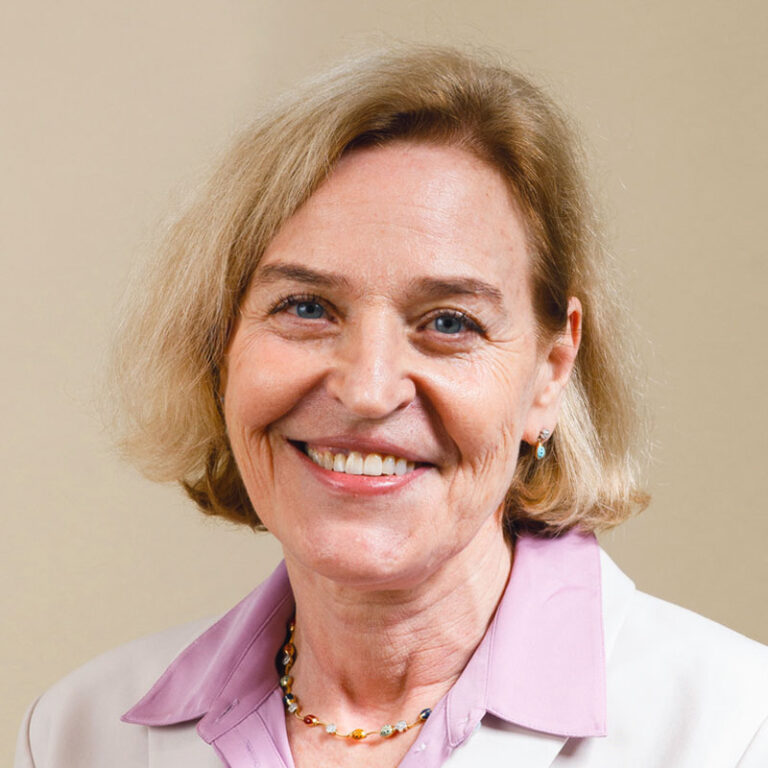 A smiling woman with shoulder-length blonde hair, wearing a white blazer over a light purple collared shirt and a beaded necklace.